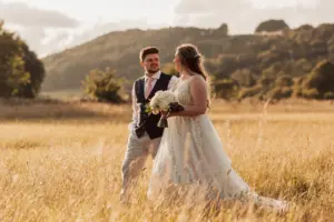 A bride and groom walk through a golden field, holding hands and gazing at each other, with rolling hills and trees in the background—an idyllic scene perfect for Eyam Hall weddings under a partly cloudy sky.