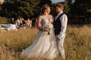 A bride in a white gown holding a bouquet stands facing a groom in a vest and tie in a sunlit field at Eyam Hall, with wedding guests and trees softly blurred in the background.