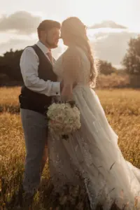 A bride and groom stand close together in a sunlit field at Eyam Hall, gazing at each other lovingly. The bride holds a bouquet of white roses, and her gown flows gently in the breeze as the sun sets behind them—perfect for romantic weddings.