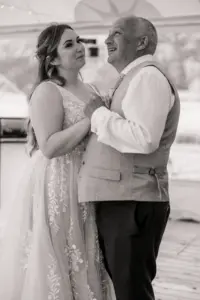 A bride in a lace wedding dress dances with an older man in a vest and tie at Eyam Hall. They hold hands and smile at each other, sharing a joyful moment in the bright, relaxed setting—capturing the happiness of weddings.