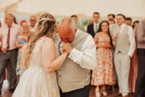 At weddings in Eyam Hall, a bride in a white lace dress shares an emotional moment as an older man, likely her father, kisses her hand. They are surrounded by smiling guests in formal attire at the elegant reception.