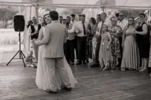 A bride and groom share their first dance at a weddings reception in Eyam Hall, surrounded by smiling guests. The scene is indoors with natural light and a wooden floor, beautifully captured in black and white.