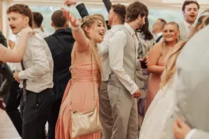 A group of people dressed formally are dancing and celebrating at an indoor weddings event, possibly at Eyam Hall; a woman in a pink dress with long hair and a gold purse smiles with arms raised among others, some holding drinks.