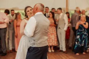 A bride in a white dress dances with an older man in a grey vest, surrounded by smiling wedding guests at Eyam Hall’s warmly lit venue.
