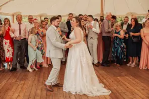 A bride and groom dance together on a wooden floor at Eyam Hall, surrounded by smiling guests in formal attire inside a decorated tent with string lights, capturing the magic of weddings.