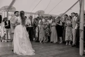 A bride and groom share their first dance in a tent at Eyam Hall, surrounded by smiling family and friends. The black-and-white photo beautifully captures the joy of weddings and this unforgettable celebration.