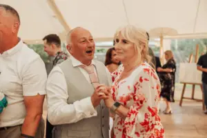 An older couple dances together, smiling and holding hands, at a lively indoor event at Eyam Hall. The man wears a light gray vest and tie, while the woman wears a white dress with red floral patterns—capturing the joy of weddings. Other guests fill the background.