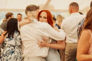 A couple embraces on a crowded dance floor at an Eyam Hall wedding. The woman with long red hair looks at the camera while holding the man, who raises a glass of white wine. People in formal attire are mingling in the background.