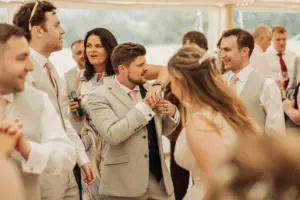 A group of people dressed in formal attire are socializing and smiling at a wedding reception at Eyam Hall. The atmosphere is lively, with guests enjoying themselves under a light-colored tent.