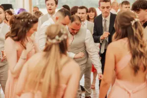A group of people in formal attire, including men in suits and women in peach dresses, dance closely together at an indoor Eyam Hall event, likely a wedding reception.