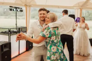 A smiling man and woman dance together inside a tent at Eyam Hall weddings, with a DJ booth and speakers nearby. In the background, a bride and another man are also dancing. The mood appears joyful and celebratory.