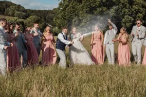 A wedding party stands in a grassy field at Eyam Hall, some wearing pink dresses and others gray suits, as a man pops a bottle of champagne, spraying it while everyone cheers and smiles under a sunny sky.