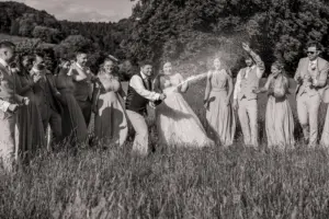 A group of people in formal attire stands in a grassy field at Eyam Hall, laughing and celebrating weddings as a man pops open a champagne bottle, spraying it into the air. The mood is joyful and festive.