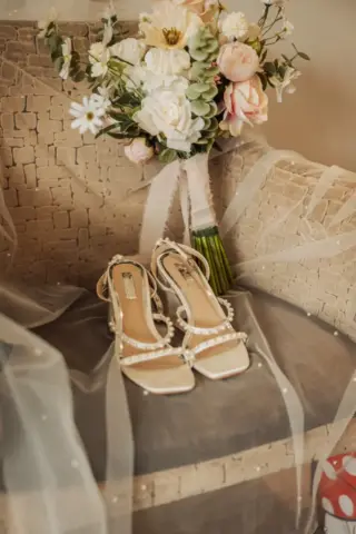 A pair of elegant, pearl-embellished bridal heels and a bouquet of white and pink flowers rest on a textured beige armchair draped with sheer, white tulle fabric—perfect details for a bride at her Whirlow Brook Hall wedding.
