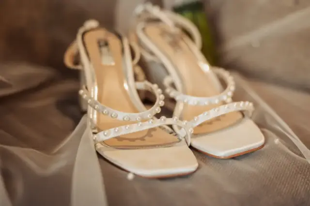 A close-up of elegant white high-heeled sandals decorated with small pearls, resting on sheer white fabric—perfect wedding shoes for a bride, possibly photographed at Whirlow Brook Hall.