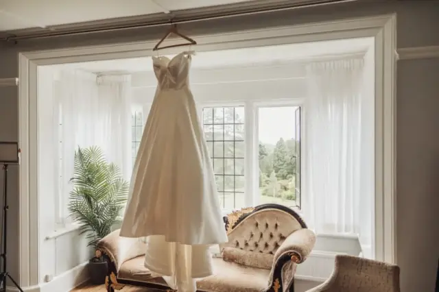 A white wedding dress hangs from a hanger in front of a large bay window with sheer curtains at Whirlow Brook Hall, next to a vintage sofa and an indoor plant, with greenery visible outside.
