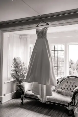A white wedding dress hangs on a hanger in front of large windows at Whirlow Brook Hall, above an ornate vintage sofa in a bright room with a potted plant and natural light streaming in.