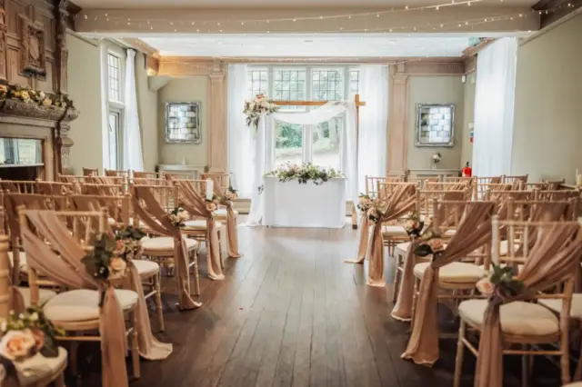 A bright, elegant wedding ceremony setup at Whirlow Brook Hall features rows of chairs draped in blush fabric and floral decorations, facing a white floral arch and table in front of tall windows with flowing white curtains.
