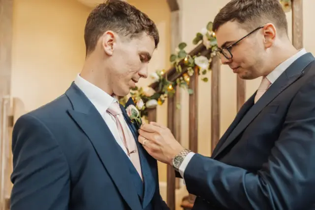 Two men in blue suits stand indoors at Whirlow Brook Hall; one adjusts the other’s boutonnière. Both wear light pink ties, with a floral garland on the railing behind them, creating a formal scene fitting for a wedding or special event.