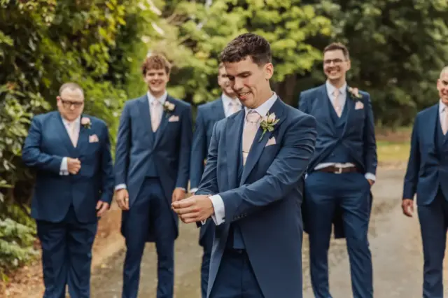 A groom in a blue suit adjusts his cufflink while smiling, surrounded by four groomsmen in matching blue suits and pink ties, standing outdoors on a tree-lined path at Whirlow Brook Hall on his wedding day.