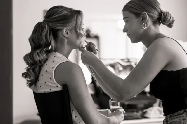 A makeup artist applies lipstick to a seated bride with wavy hair, who holds a glass and looks ahead. The softly lit room at Whirlow Brook Hall is captured in black and white, adding timeless elegance to this wedding moment.