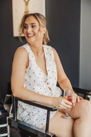A smiling bride in a white, sleeveless dress with small red hearts sits on a chair at Whirlow Brook Hall, holding a glass of pink sparkling drink. Her hair is tied back, and she appears relaxed and happy in the modern indoor setting.