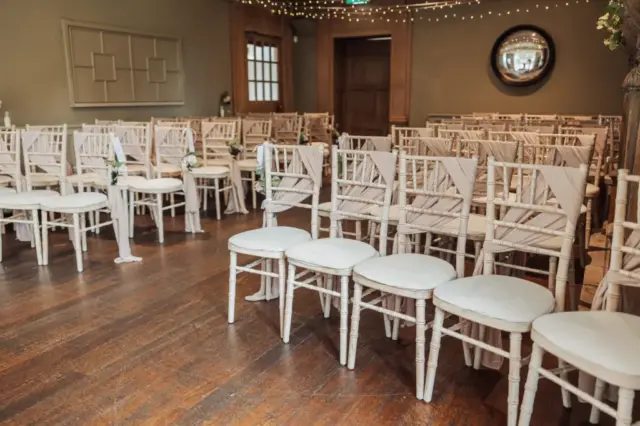 Rows of white chairs with light fabric draping are arranged for a wedding ceremony at Whirlow Brook Hall, an elegant room with wooden floors, beige walls, string lights, and a round mirror—perfect for the bride’s unforgettable moment.
