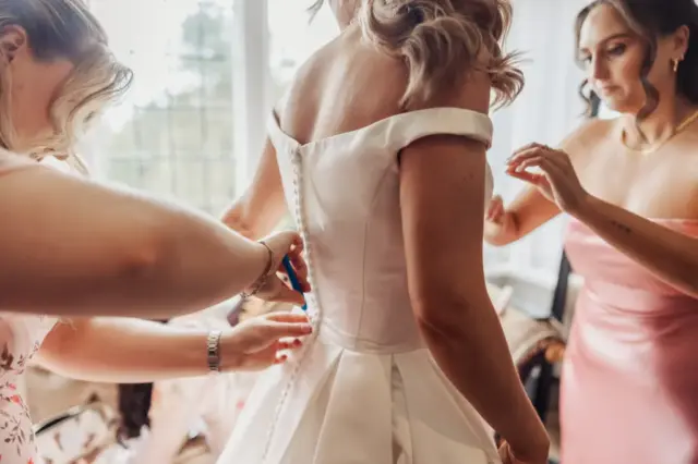 Three women help a bride button up her white, off-the-shoulder wedding dress at Whirlow Brook Hall. The scene is indoors with natural light streaming through a window in the background.