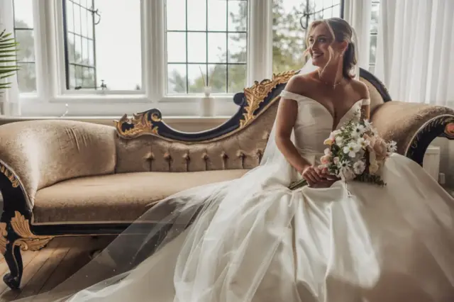 A bride in an off-the-shoulder white wedding gown sits on an ornate vintage sofa at Whirlow Brook Hall, holding a bouquet and smiling, with large windows and soft natural light in the background.