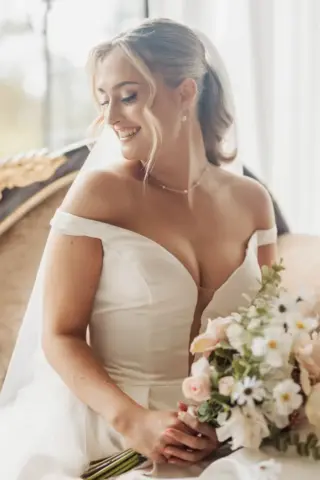 A smiling bride in an off-the-shoulder white gown holds a bouquet of blush flowers, sitting on a vintage sofa at Whirlow Brook Hall, with soft natural light streaming in through the window behind her.