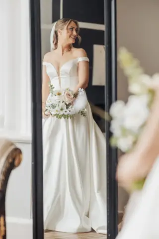 A smiling bride in an off-shoulder white wedding gown stands before a mirror at Whirlow Brook Hall, holding a bouquet of white and blush flowers, her reflection clearly visible.