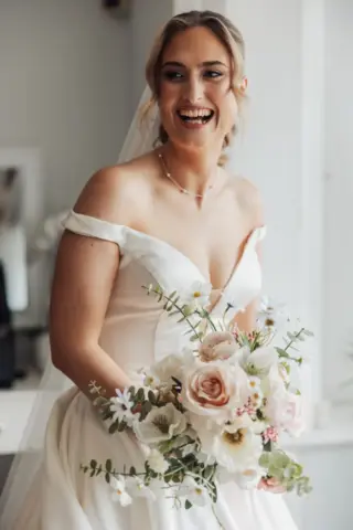 A smiling bride in an off-the-shoulder white wedding dress holds a bouquet of pale pink and white flowers at Whirlow Brook Hall. She stands indoors, her hair styled back, wearing a delicate necklace and veil.