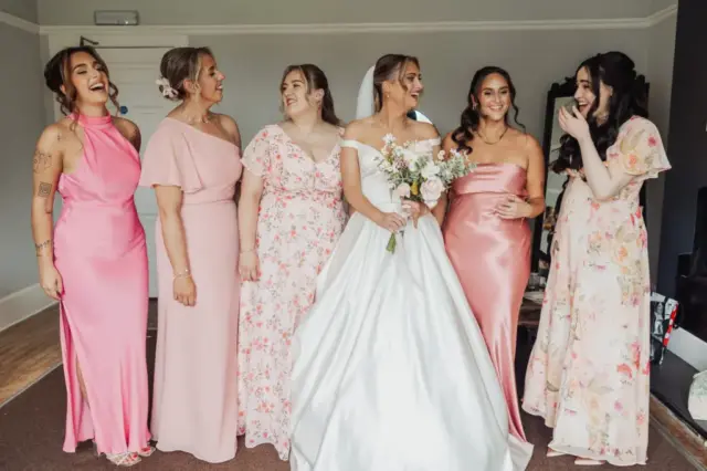 Six women stand indoors, smiling and laughing together. The bride in a white wedding dress holds a bouquet, while her bridal party in pink or floral dresses suggests they’re preparing for a wedding at Whirlow Brook Hall.