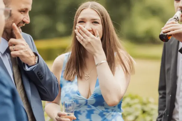 A young bride in a blue dress laughs and covers her mouth with her hand while holding a drink, surrounded by people in suits at a Whirlow Brook Hall wedding outdoor gathering.