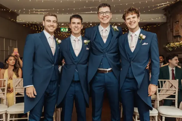 Four men in matching blue suits with pale pink ties and boutonnieres stand smiling together indoors at a Whirlow Brook Hall wedding, with guests seated in the background and string lights overhead.
