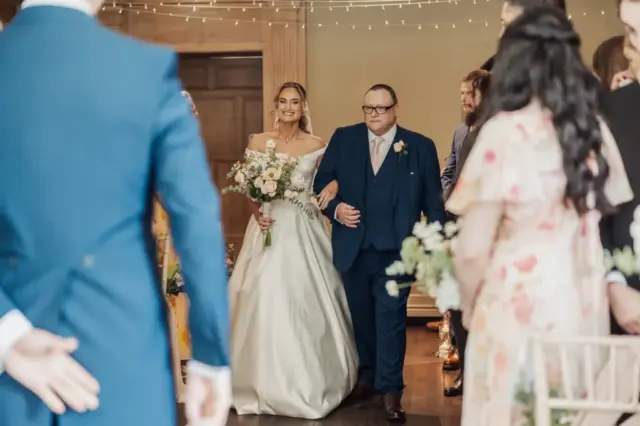 A smiling bride in a white dress walks down the aisle at Whirlow Brook Hall, holding a bouquet and accompanied by a man in a blue suit, as guests watch and the groom waits beneath glowing string lights.