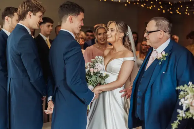 A bride and groom stand smiling and holding hands during their wedding ceremony at Whirlow Brook Hall, surrounded by guests and groomsmen. The bride holds a bouquet as an older man in a blue suit stands beside them, speaking.