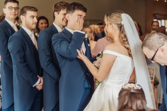 A groom wipes away tears while facing his bride during their Whirlow Brook Hall wedding ceremony. The bride holds his hand as groomsmen in blue suits smile behind them, and guests watch the emotional moment.