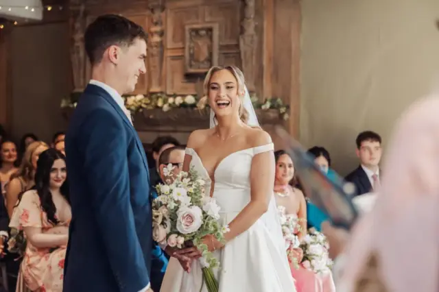 A bride in a white dress and a groom in a blue suit smile at each other during their wedding ceremony at Whirlow Brook Hall, with guests seated and watching in the background. The bride holds a bouquet of flowers.