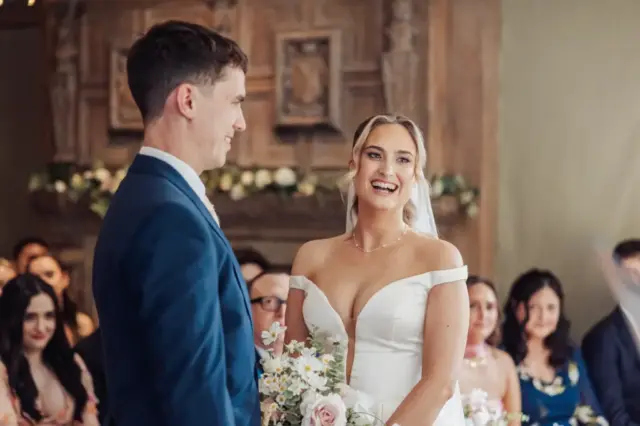 A bride in a white dress and veil smiles joyfully at the groom in a blue suit during their Whirlow Brook Hall wedding ceremony, with guests seated and watching in the background.