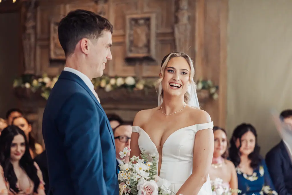 A bride in a white dress and veil smiles joyfully at the groom in a blue suit during their Whirlow Brook Hall wedding ceremony, with guests seated and watching in the background.