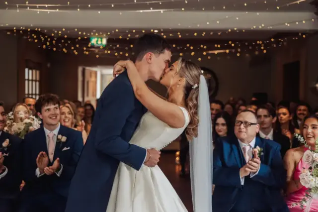A bride and groom share a kiss at their indoor Whirlow Brook Hall wedding ceremony, surrounded by guests who are smiling and clapping under strings of twinkling fairy lights.