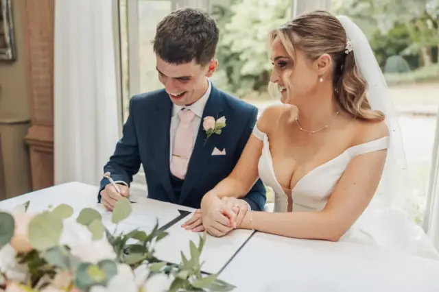 A bride and groom sit at a table, smiling and holding hands, as the groom signs a document during their wedding at Whirlow Brook Hall. The bride wears an off-shoulder white gown and veil; the groom is in a navy suit with a pale pink tie and boutonniere.