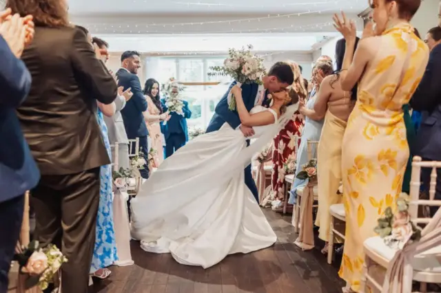 A bride and groom share a kiss as the groom dips the bride, surrounded by applauding guests in colorful outfits during a beautiful wedding at Whirlow Brook Hall.