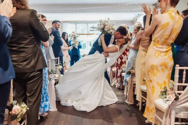 A groom dips and kisses his bride as they stand at the end of an aisle at Whirlow Brook Hall, surrounded by applauding guests in colorful attire during an indoor wedding ceremony.