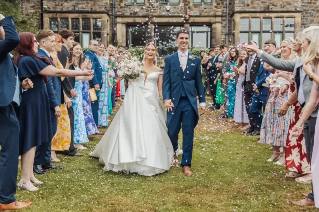 A bride and groom walk hand-in-hand outside Whirlow Brook Hall, smiling as guests on both sides throw confetti. The bride wears a white gown and holds a bouquet; the groom is in a navy suit. Wedding guests cheer in colorful outfits.