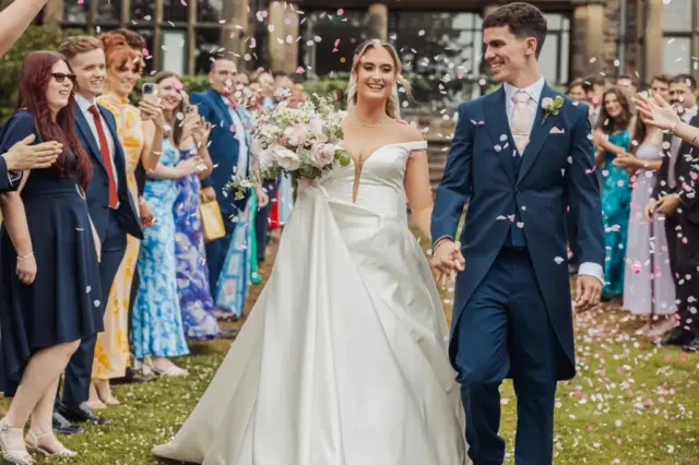 A smiling bride and groom walk hand in hand outdoors at Whirlow Brook Hall as guests in colorful attire throw pink and white confetti, celebrating their wedding. The bride holds a bouquet and wears a white dress; the groom wears a dark suit.