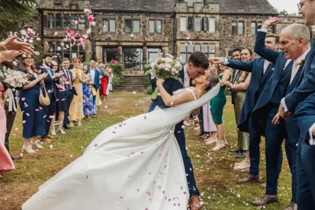 A bride and groom kiss in a joyful dip outside Whirlow Brook Hall as guests throw pink and white flower petals. The wedding scene is festive, with smiling friends and family cheering them on.