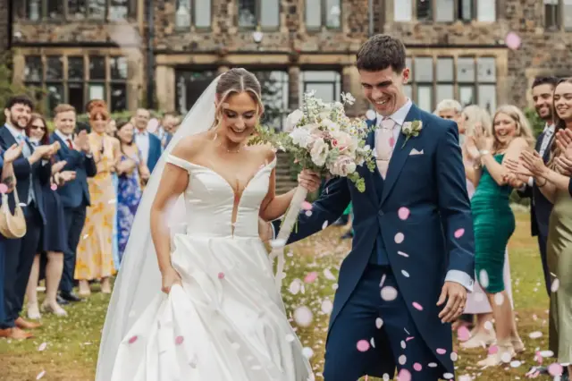 A bride and groom smile and walk outdoors at Whirlow Brook Hall while guests throw pink and white confetti. The bride wears a white dress and veil; the groom is in a navy suit as everyone celebrates in front of the grand stone building.