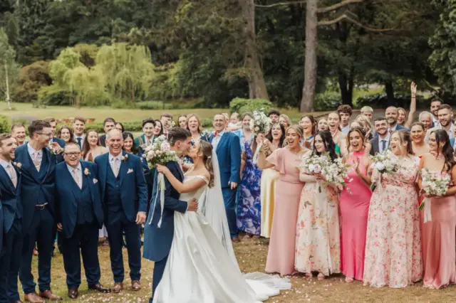 A bride and groom kiss outdoors at Whirlow Brook Hall, surrounded by their wedding party and guests in colorful dresses and suits. The group smiles and celebrates amid lush green trees and gardens.